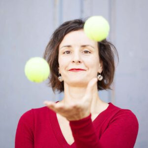Junge Frau im roten Pullover jongliert mit zwei Tennisbällen vor hellblauer Wand. Sie konzemtriet sich auf die Bälle.