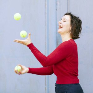 Junge Frau im roten Pullover jongliert mit drei Tennisbällen vor hellblauer Wand. Dabei lacht die Frau.