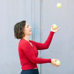 Junge Frau im roten Pullover jongliert mit drei Tennisbällen vor hellblauer Wand.