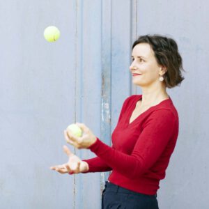 Junge Frau im roten Pullover jongliert mit Tennisbällen vor hellblauer Wand